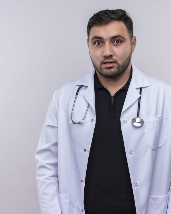 young bearded male doctor wearing white coat with stethoscope looking at camera confused standing over wite background